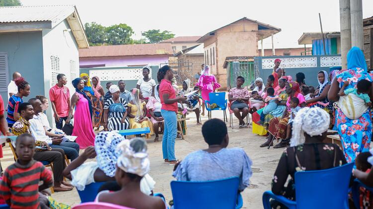 Eine Gesundheitsberaterin im roten T-Shirt des KCCR steht in der Mitte eines Kreises von etwa 50 stehenden und sitzenden Frauen, Männern und Kindern auf einem Platz umgeben von einigen Häusern.